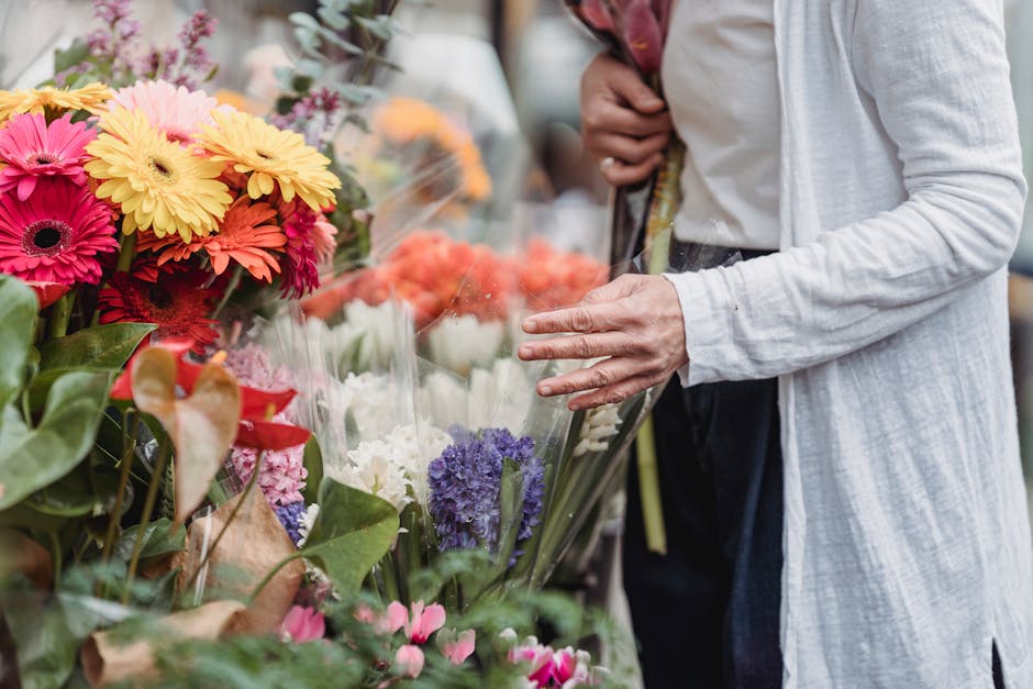 A person wearing a light grey, long-sleeved linen jacket is selecting a bouquet of fresh flowers at a flower shop on Silk Street EC2Y. The bouquet includes bright pink and yellow gerbera daisies, purp
